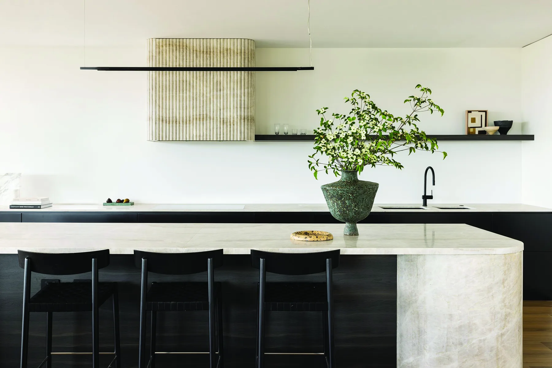 Mosman &mdash; kitchen island in pale stone with a fluted rangehood and black stools